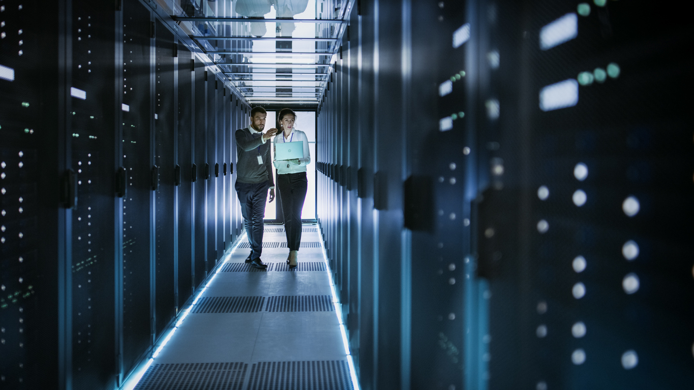 Female IT Technician and Male Server Engineer Talk and Discuss. They are in Working Data Center full of Rack Servers. Woman Holds Laptop.